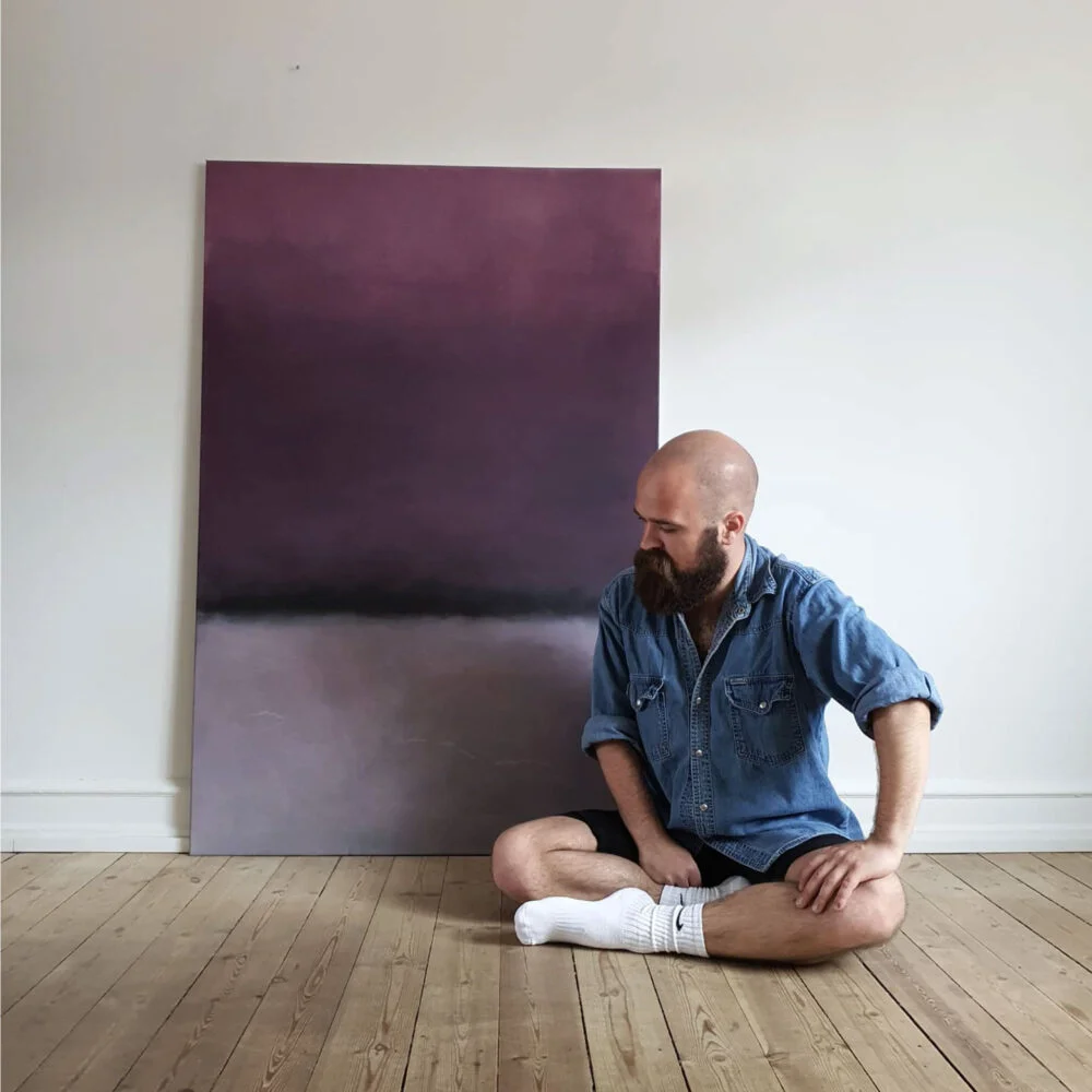 Frederik Smal seated on the floor beside a large abstract painting in deep purple and grey